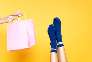 Cropped view of woman holding pink paper bags near female legs in blue socks isolated on yellow background