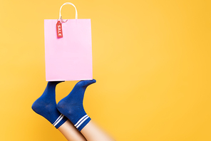 Top view of female legs in blue socks holding paper bag with sale label isolated on yellow background