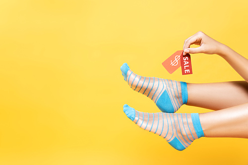 Cropped view of woman wearing striped socks, holding tags with dollar symbol and sale word isolated on yellow