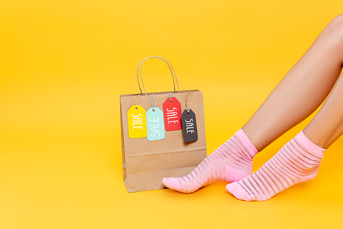 Cropped view of female legs in socks with pink stripes near paper bag with tags with sale words isolated on yellow