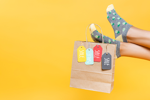 Cropped view of paper bag with sale tags hanging on woman legs in socks isolated on yellow