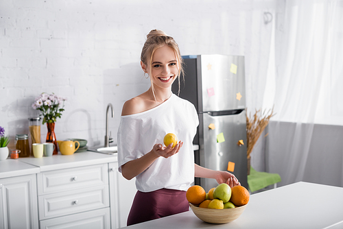 smiling blonde woman holding lemon while standing near bowl with fresh fruits