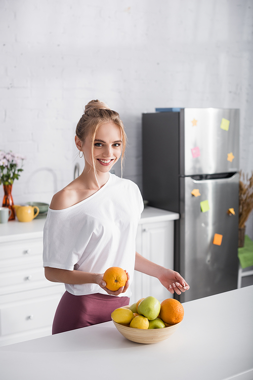 happy woman in white t-shirt smiling at camera while holding ripe orange near bowl with fresh fruits