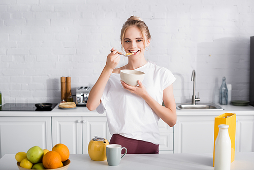 happy blonde woman eating cornflakes near table with fruits, teapot, and bottle of milk