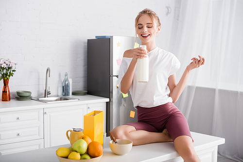 cheerful blonde woman holding bottle of milk while sitting on kitchen table near fruits