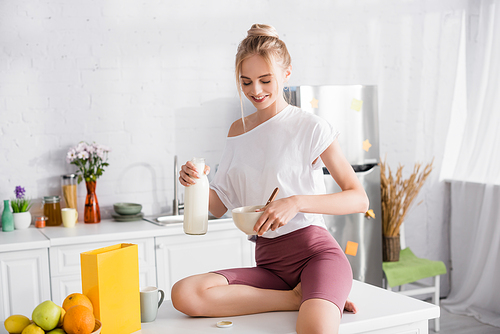 smiling blonde woman sitting on kitchen table and pouring milk into bowl