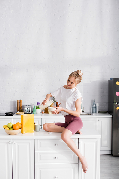 barefoot woman in shorts pouring milk into bowl while sitting on table in kitchen