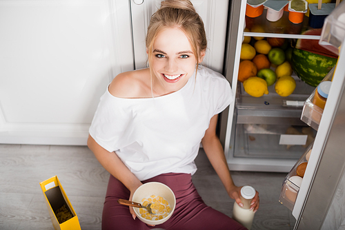 blonde woman sitting on floor near opened fridge with bowl of cornflakes and bottle of milk