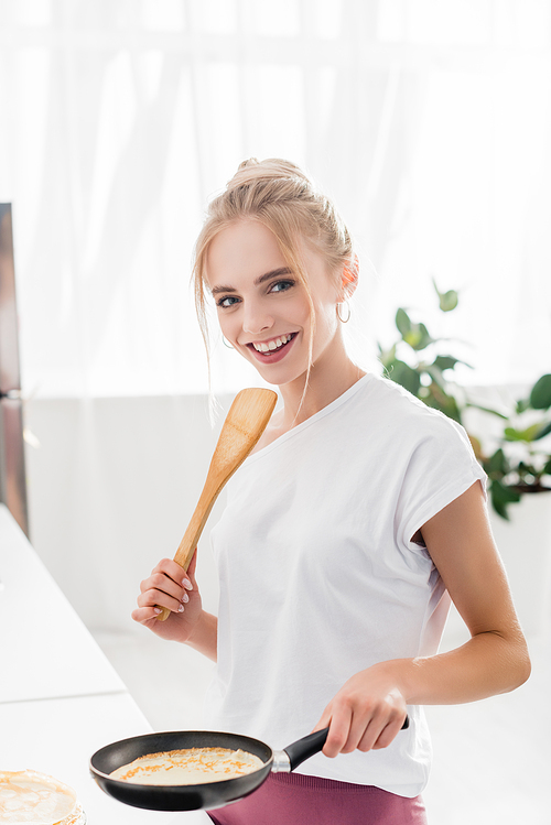 blonde woman in white t-shirt holding spatula and frying pan with tasty pancake