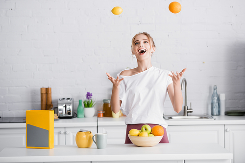 excited young woman juggling with fresh fruits while standing near teapot and cup