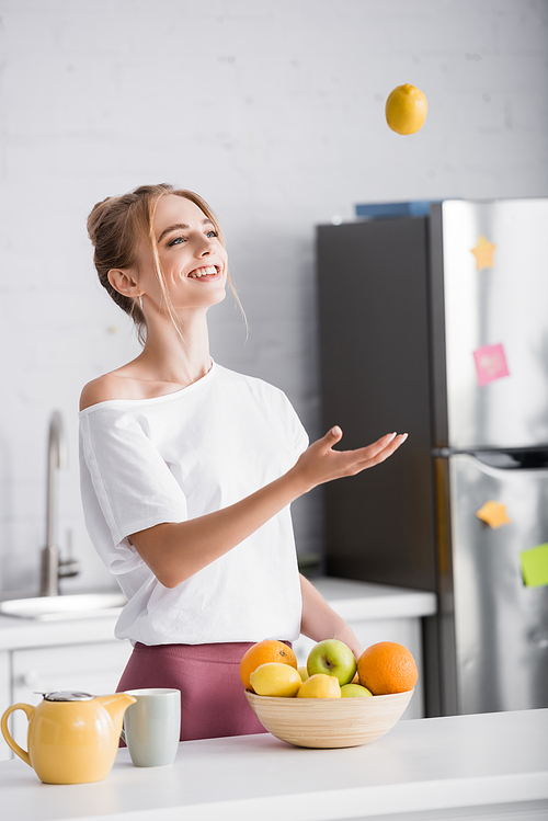 joyful young woman juggling with ripe lemon while standing near teapot and cup