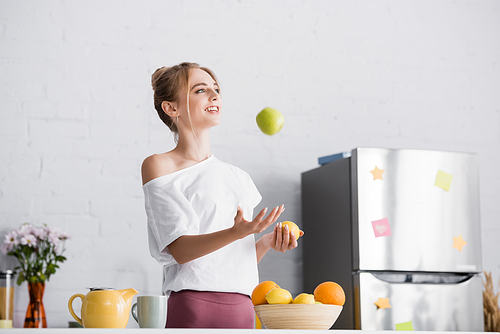 young woman in white t-shirt juggling with fresh fruits near teapot and cup in kitchen