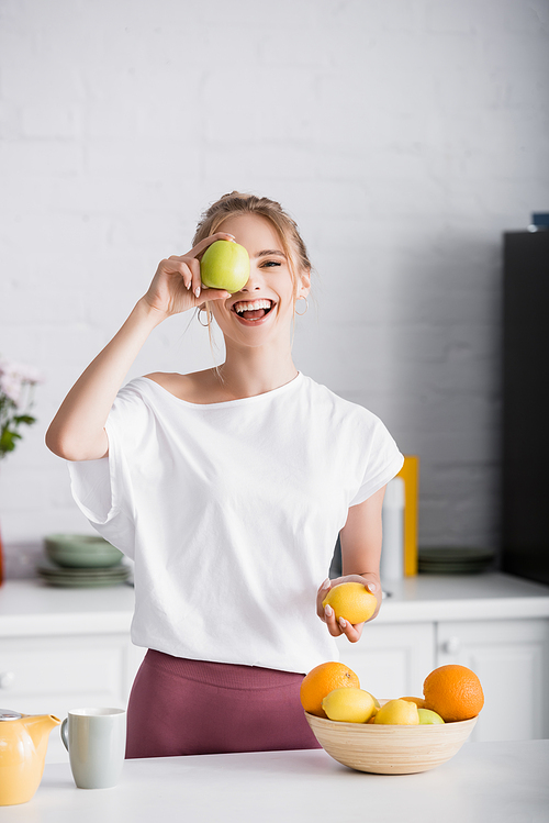 cheerful young woman covering eye with apple while holding lemon in kitchen