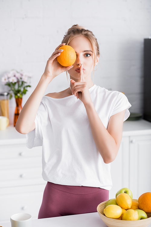 young blonde woman obscuring eye with fresh orange and showing hush gesture