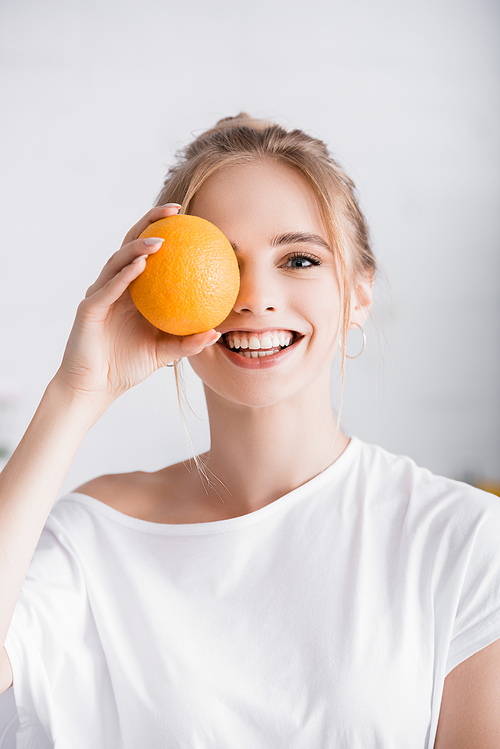 happy blonde woman  while covering eye with whole ripe orange
