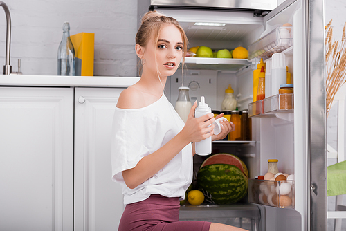 sensual woman in white t-shirt holding bottle of whipped cream while sitting near open fridge