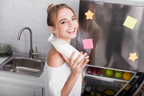 happy blonde woman smiling at camera while holding bottle of milk near fridge