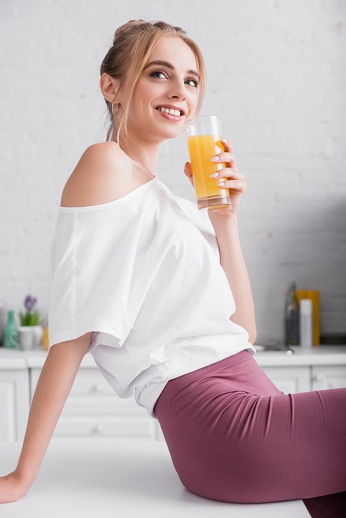 smiling blonde woman in white t-shirt holding glass of orange juice while sitting on kitchen table