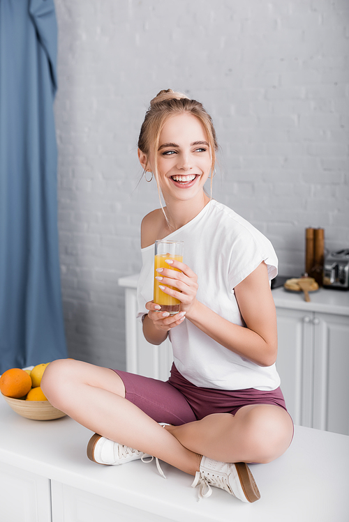 happy young woman in white t-shirt sitting on kitchen table with crossed legs and holding orange juice