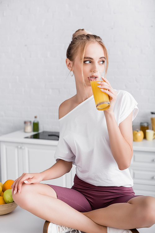 blonde woman sitting on kitchen table with crossed legs and drinking orange juice