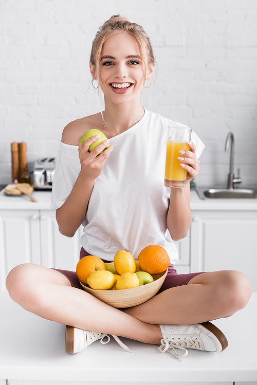 pleased young woman smiling at camera while sitting on kitchen table with fruits and glass of orange juice