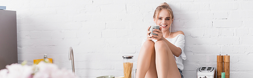 selective focus of joyful woman holding cup while sitting on kitchen table, website header