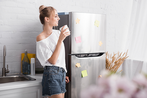 selective focus of blonde woman in white t-shirt and denim shorts standing in kitchen with cup of tea
