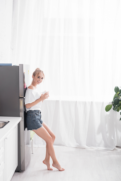 young blonde woman in denim shorts standing near fridge with cup of tea and 