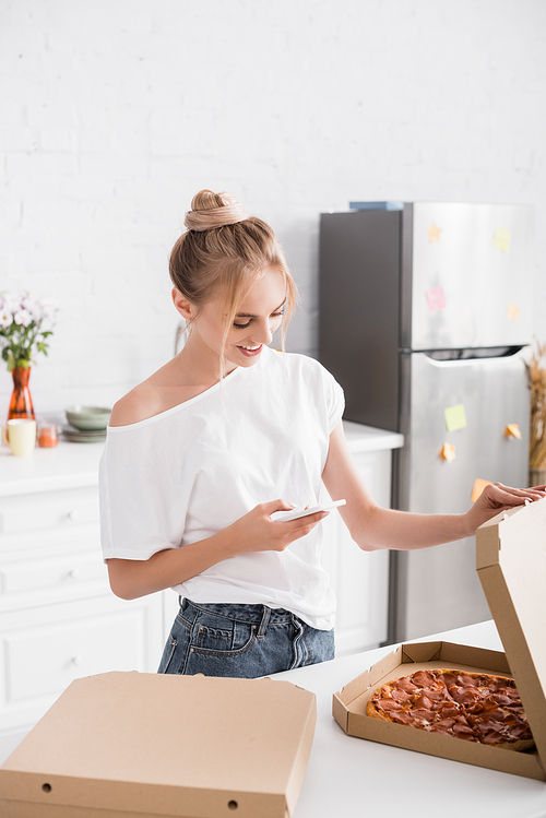 young woman in white t-shirt messaging on mobile phone while opening box with pizza in kitchen