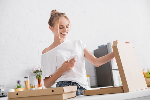 pleased young woman messaging on smartphone while opening box with pizza