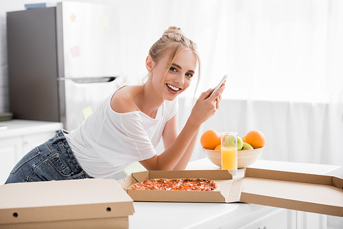 excited young woman chatting on smartphone near pizza and fresh fruits in kitchen