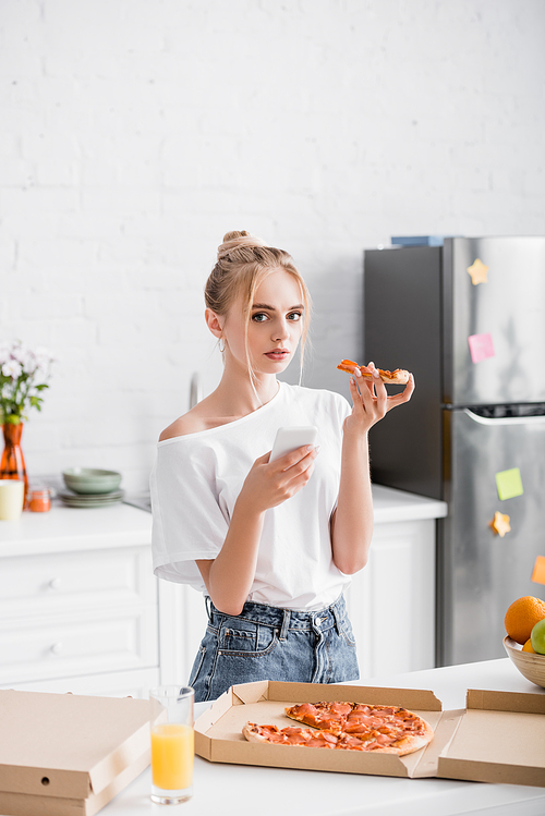 blonde woman holding smartphone and pizza while  in kitchen