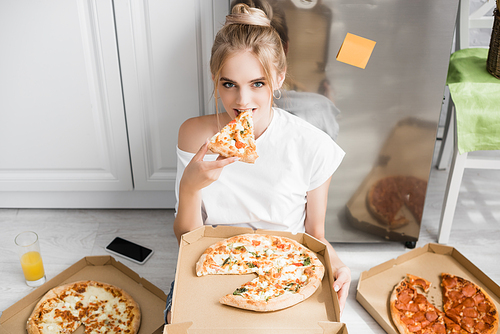 young blonde woman  while sitting on floor in kitchen and eating pizza