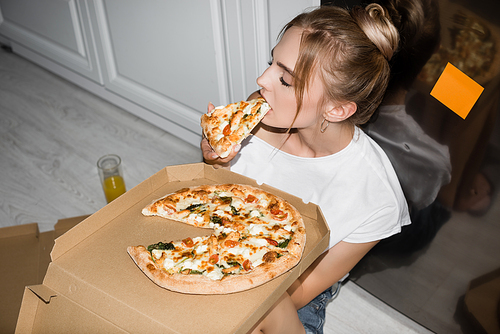 high angle view of young blonde woman eating pizza while sitting on floor in kitchen