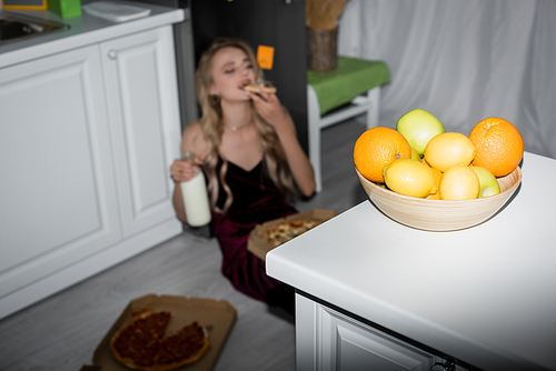 selective focus of young woman eating pizza and holding milk while sitting on floor near table with fresh fruits
