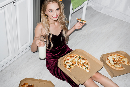 high angle view of cheerful woman in velour dress eating pizza and holding bottle of milk while 