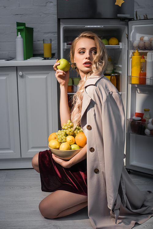 blonde woman in trench coat holding bowl with fresh fruits near opened fridge