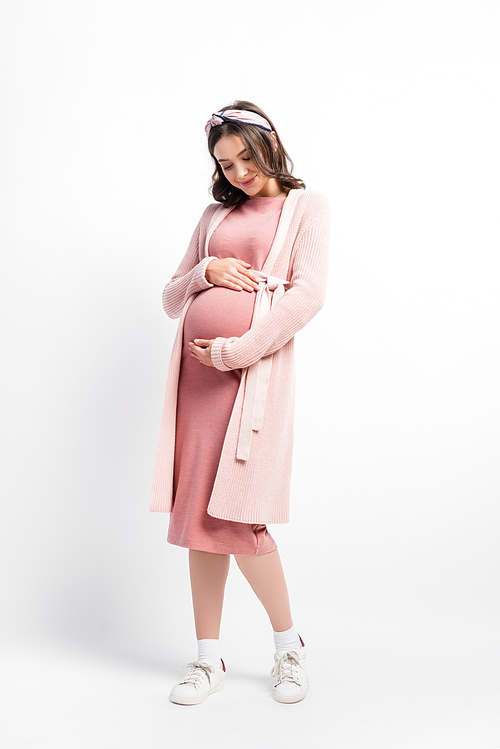 full length view of pregnant woman in cardigan and headband touching belly on white