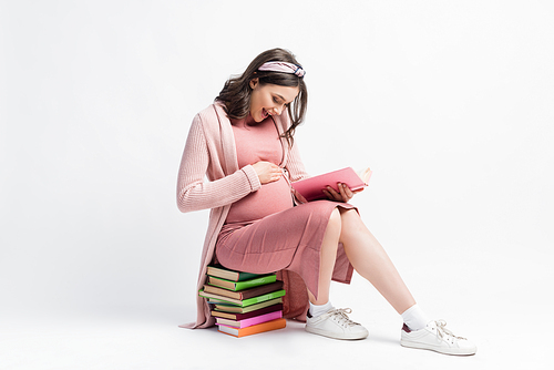 excited pregnant woman sitting on books and touching belly on white