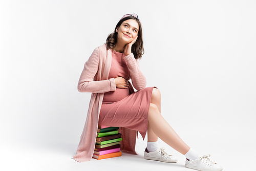 dreamy and pregnant woman sitting on books and looking away on white