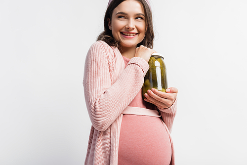 joyful and pregnant woman holding jar with pickled cucumbers isolated on white