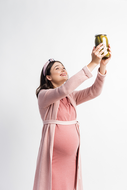 pleased and pregnant woman holding jar with pickled cucumbers above head isolated on white