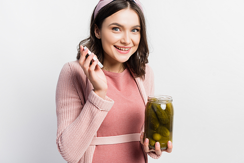 joyful and pregnant woman holding jar with pickled cucumbers isolated on white