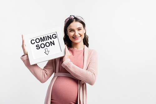 pleased and pregnant woman holding board with coming soon lettering isolated on white