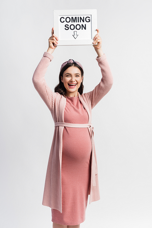 excited and pregnant woman holding board with coming soon lettering isolated on white