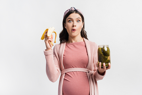 pregnant woman eating banana while holding jar with canned cucumbers isolated on white