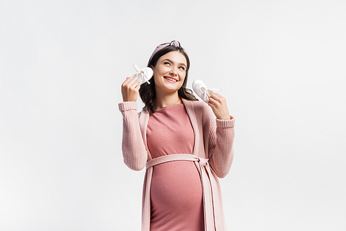 joyful and pregnant woman holding tiny boots and looking up isolated on white