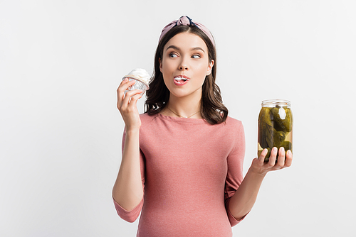 pregnant woman eating cupcake and holding jar with pickled cucumbers isolated on white