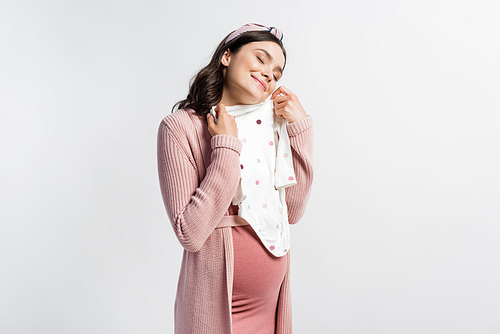 joyful and pregnant woman in headband holding baby romper isolated on white