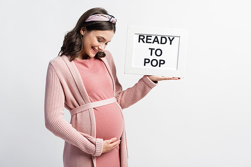 pleased pregnant woman holding board with ready to pop lettering isolated on white